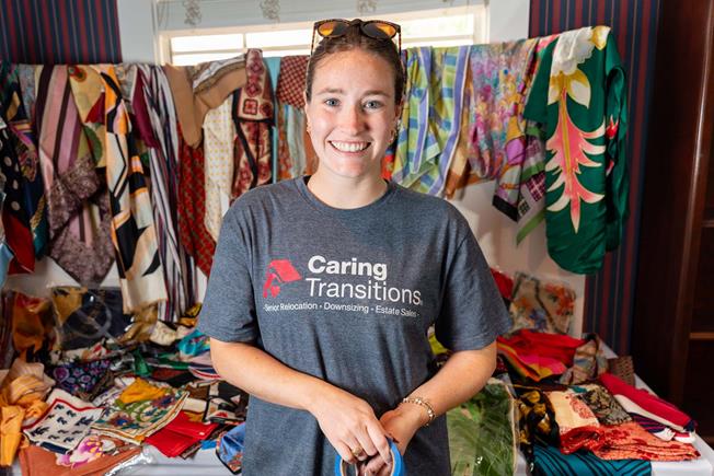 Smiling women in a 'Caring Transitions' t-shirt stands in front of a colorful scarves displayed on a table.